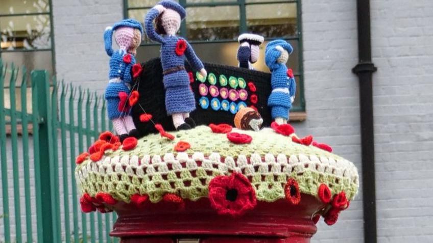 A knitted WW2 Bombe machine with Women's Royal Naval Service operators in uniform, saluting, sits atop a traditional red post box, outside a grey brick building with a green fence.