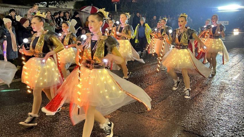 Young girls in outfits covered in sparkles and fairy lights dance down a street in Glastonbury as part of their carnival. 