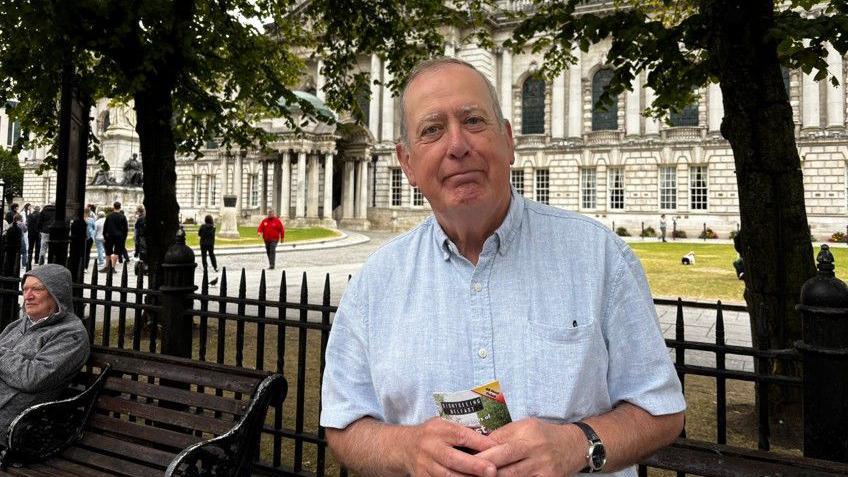A man, Hugh O'Donnell. Pictured outside Belfast City Hall, he is wearing a blue button up shirt with a black watch on his left wrist. He is smiling with a booklet in his hand.