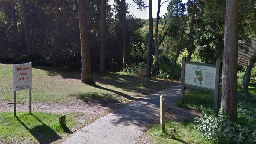 A woodland with grass and trees. A sign with a map says Lickey Hills Country Another sign reads this gate closes at dusk. 

