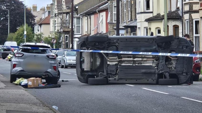 An image showing the underside of car on its side next to a grey car. Both are stationary in a residential road in Lowestoft. Police tape can be seen cordoning off the scene.