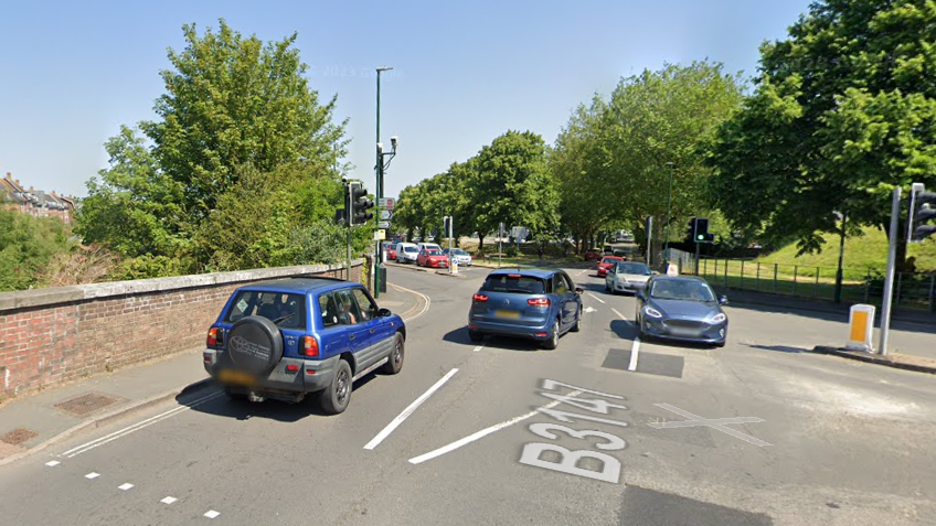 Cars driving on the four-way junction at Maumbury Cross in Dorchester. To the left is a brick wall with a narrow pavement.