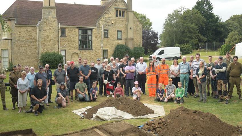 Groups shot of participants and visitors at the dig site standing in front of Fulbeck Manor with mounds of earth in the foreground