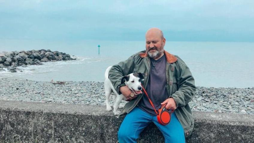Mr Stanik sitting on a wall on a sea front with a shale beach and sea behind him. next to him is a jack russell terrier