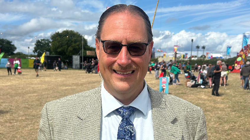 Martin smiling looking at the camera. He is standing on the eisteddfod field with lots of people walking in the background. Martin is wearing a beige suit, a light blue shirt, a patterned navy tie and black sunglasses. It is a head and shoulders shot of him.