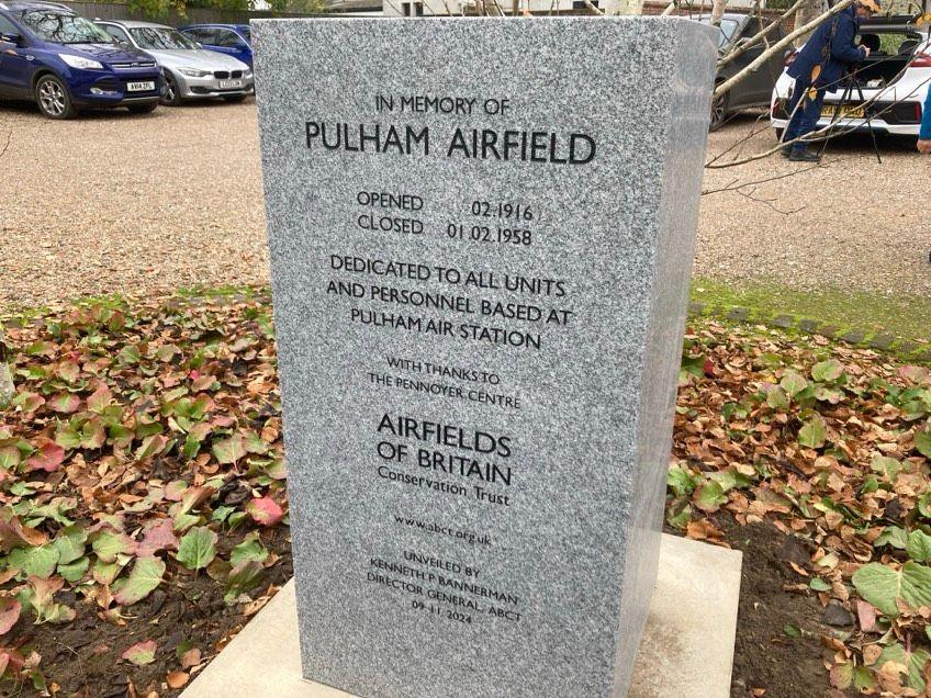 The Pulham airfield grey memorial sits on small patch of grass. It reads, 'In memory of Pulham airfield' with the opening and closing dates underneath. Cars can be seen parked behind it on a gravel surface.