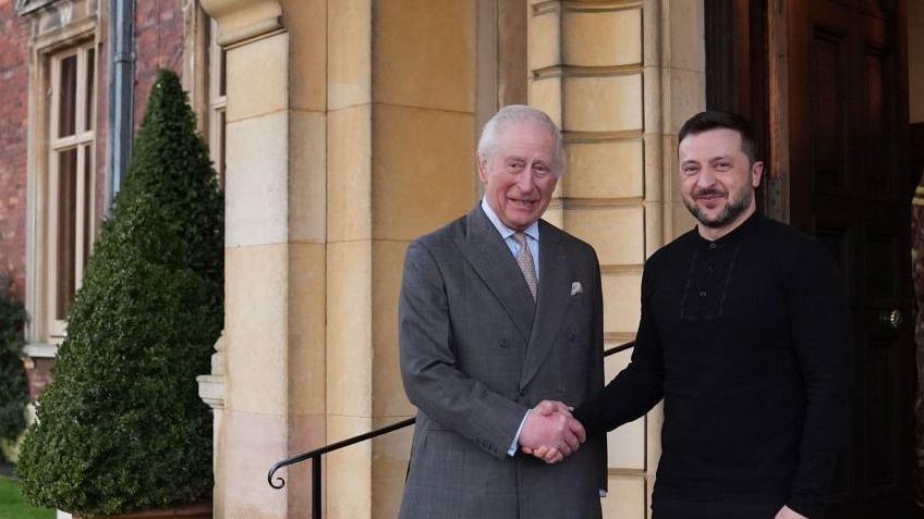 King Charles with Volodymyr Zelensky, president of Ukraine. They are shaking hands and smiling on the steps of Sandringham House