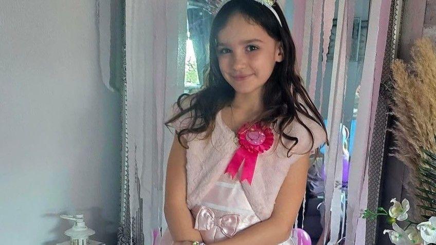 Aria Thorpe wearing a pink party dress with a pink birthday badge and a tiara. She is standing in a living room on her birthday, with silver and pink streamers hanging from a doorway behind her. 