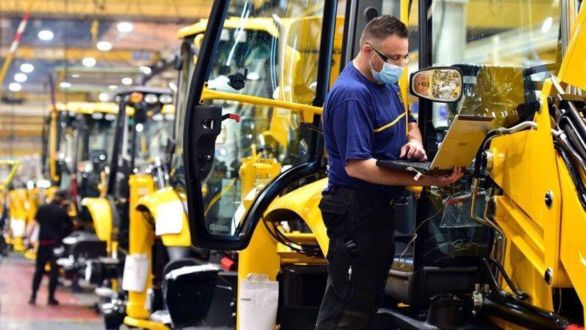 On a factory production line, a man in a blue t-shirt and wearing a medical mask examines an open laptop as he stands beside a partially-constructed yellow JCB digger.