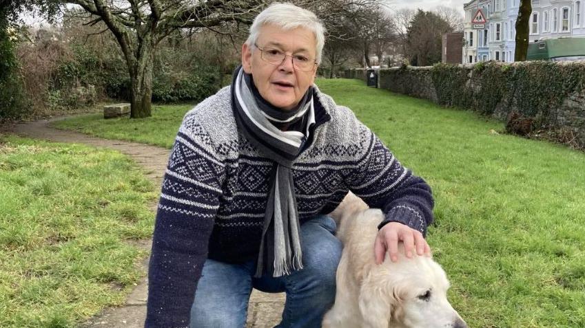 Chris Hockaday wearing a blue and white nordic style jumper and a black, grey and white scarf and blue jeans kneels down in a park. He is stroking his golden retriever dog.