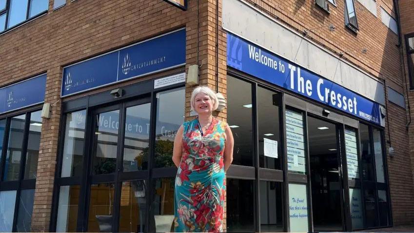 Ms Chantal is standing outside the Cresset building. It has brown bricks and electric glass doors at the entrance. She is smiling with her hands behind her back.