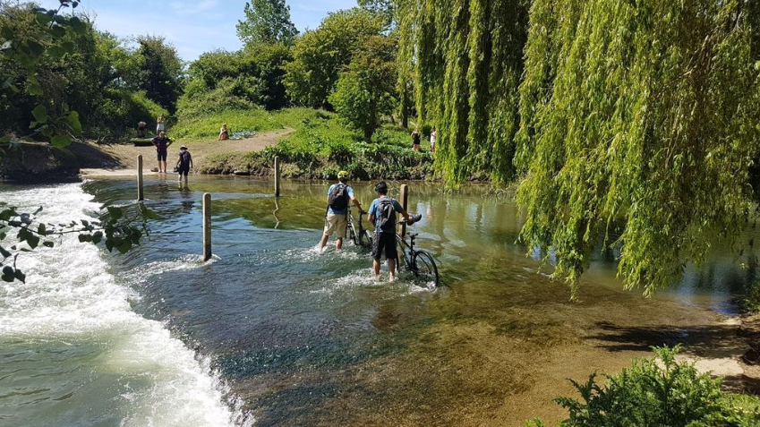 Two people pushing their bicycles through shallow water at Duxford in Oxfordshire.