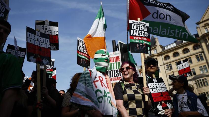 A crowd of people holding placards saying 'Defend Kneecap', and holding Palestinian flags and Irish flags outside the court in London