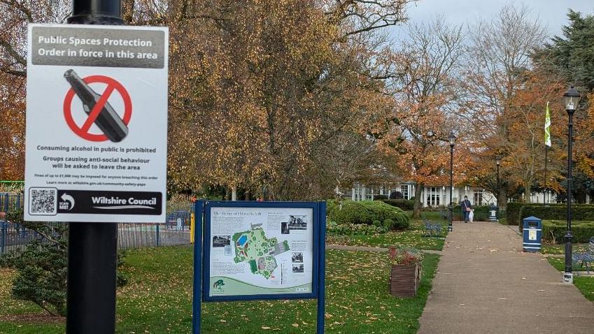 A scene in a Salisbury park showing a Public Spaces Protection Order poster. In the background is an autumnal park scene. 