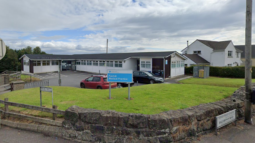 A Google Maps screenshot of Fairhill Medical Practice. The image shows a single storey white building with columns of dark brick on its exterior. In front of the building is a car park and a blue and white sign reading 'Fairhill Medical Practice'.