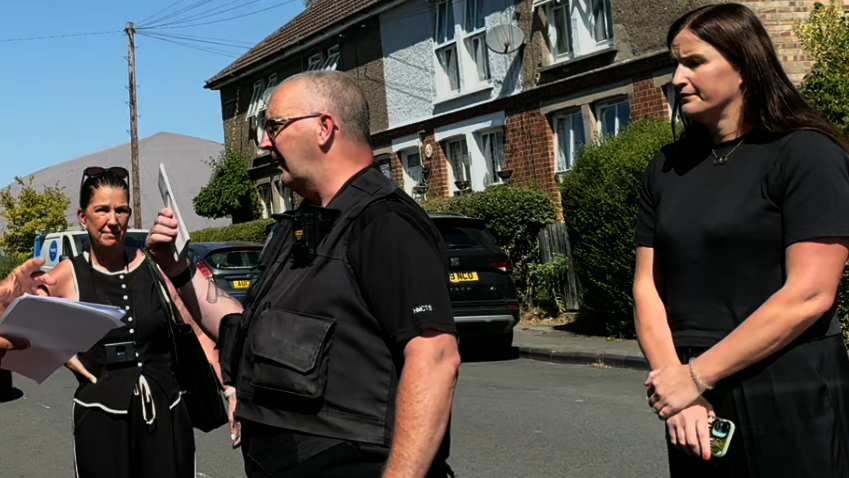 Two women dressed in black standing either side of a male bailiff, who is also dressed in black and holding up a piece of paper.