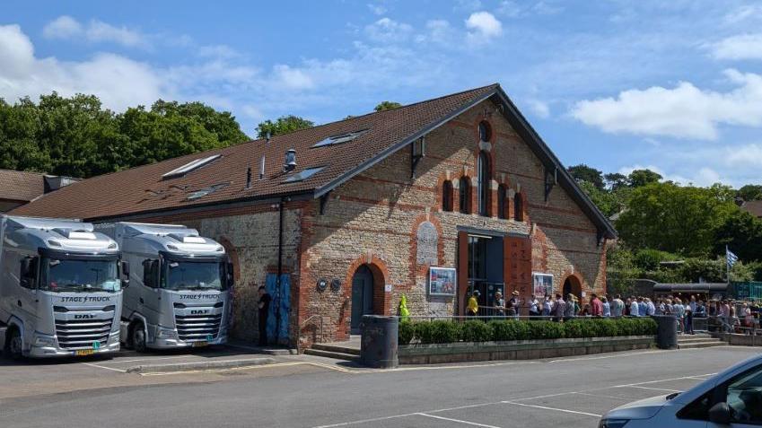 A crowd of people standing outside the Cheese and Grain venue in Frome on a bright, blue sky day. There are large tour trucks parked next to the stone building. 