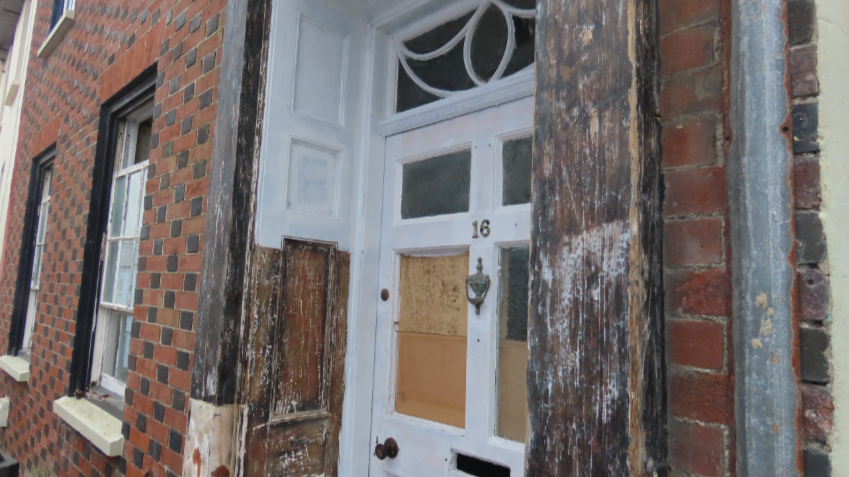 A close-up of a boarded up door on the street. Around the front door is rotting wood.