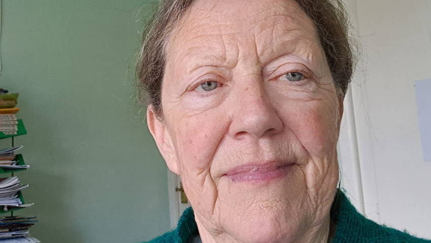 A picture of an older woman looking at the camera against a green wall.
