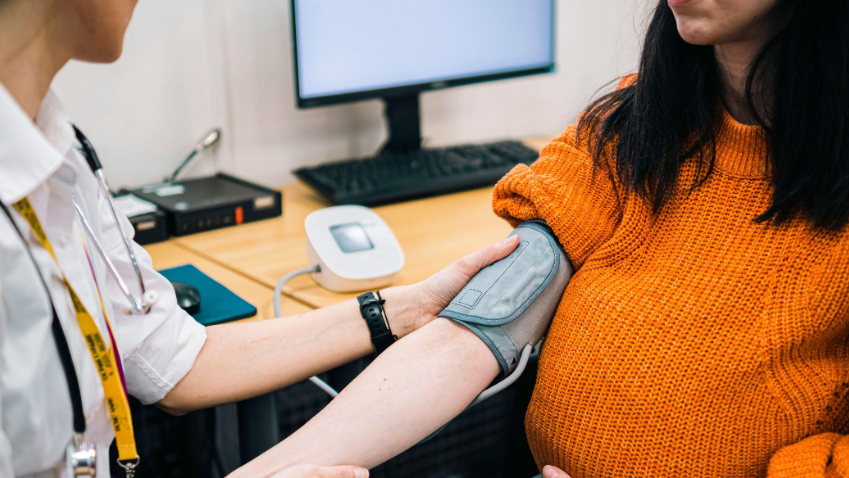 A doctor checking the blood pressure of a patient. Their faces are not in the picture. The male doctor is wearing a white shirt and the female patient is wearing a bright orange jumper