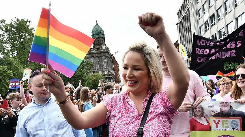 Michelle O'Neill has blonde hair and is wearing a pink t-shirt. She is smiling at the crowd and waving a rainbow Pride flag and pumping her fist while walking among lots of people at a Pride parade in Belfast