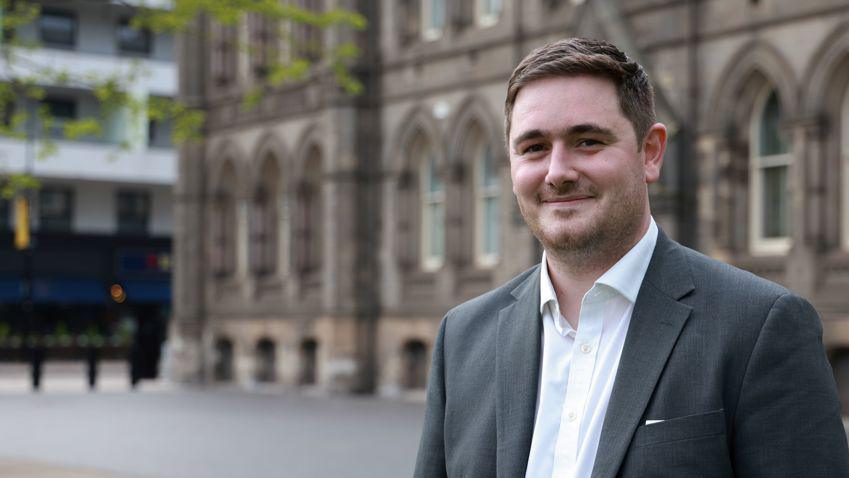 Man in grey suit and white shirt with top button undone, smiling in front of an ornate building