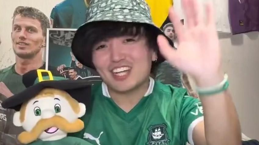 A man in a green outfit and bucket hat waves and holds a Plymouth Argyle mascot.