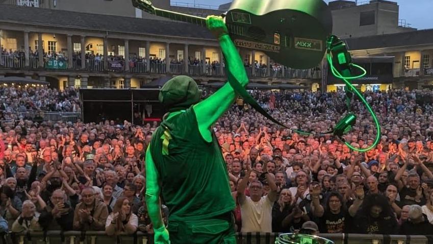 Mike Peters on stage at a gig holding a guitar, with his back to the camera and a massive outdoor crowd facing the stage