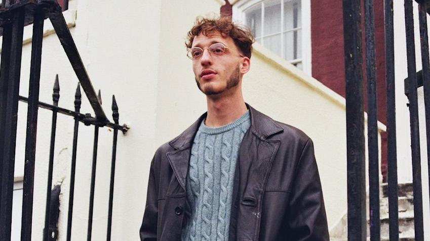 Press shot of Elijah A.M in a blue jumper and coat standing near some railings