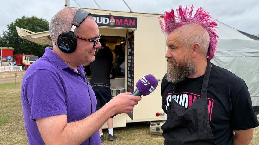 Chris Goreham wearing a purple BBC Radio Norfolk polo shirt holding a microphone and he is interviewing Spud man. Spud man, also known as Ben Newman, is wearing a black T-shirt with pink hair. 