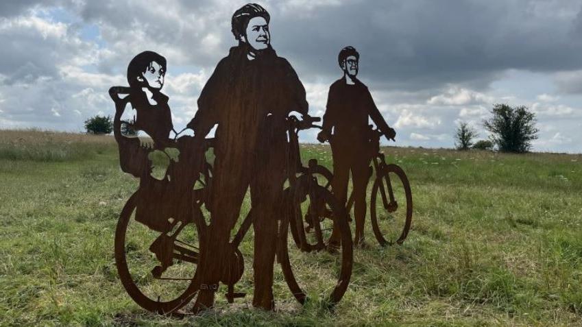 Steel statues of silhouettes on Salisbury Plain of a woman wearing a bicycle helmet holding a bicycle with a small child in a bike seat. A man holding a bicycle is in the background.