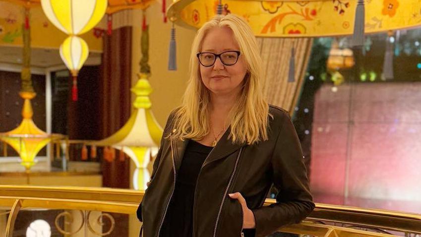 A woman with long blonde hair and black-rimmed glasses stands in front of a railing with lights and bright decorations in the background. She is wearing a black top underneath what looks like a black leather jacket.