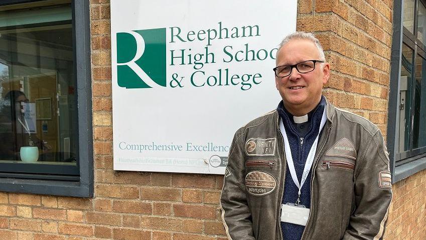 Reverend Keith Rengert stands outside Reepham High School sign wearing a dog collar under his leather jacket