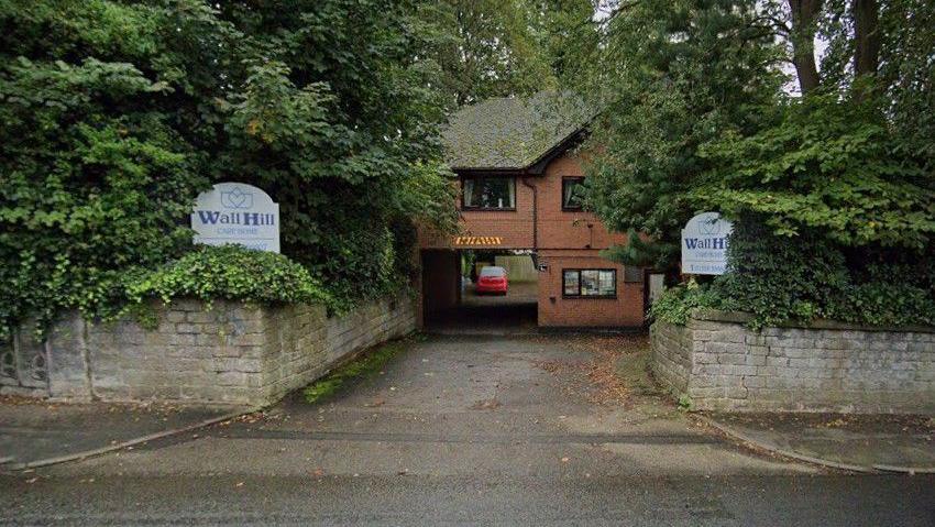 The entrance to a care home, with stone walls either side. Signs either side of the road say Wall Hill Care Home in blue lettering on a white background.