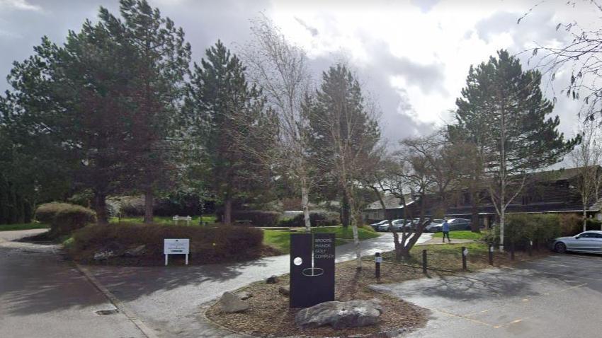 Front of the golf complex, with sign, trees, parked cars and clubhouse building in the background