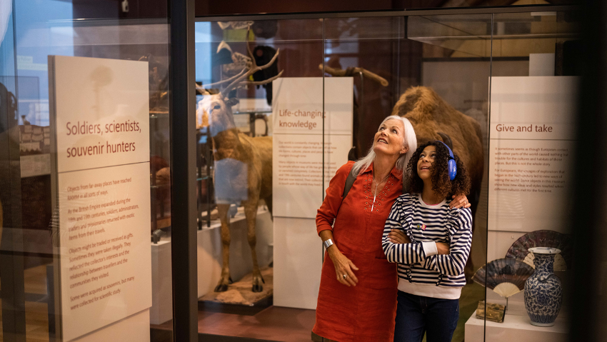 A woman, with white blonde hair and wearing a red dress, and a younger girl, wearing blue headphones and stripy jumper and black trousers, both looking up. Behind them are exhibits with a animals in them and to the right are a vase and two fans.  There are information boards around them.