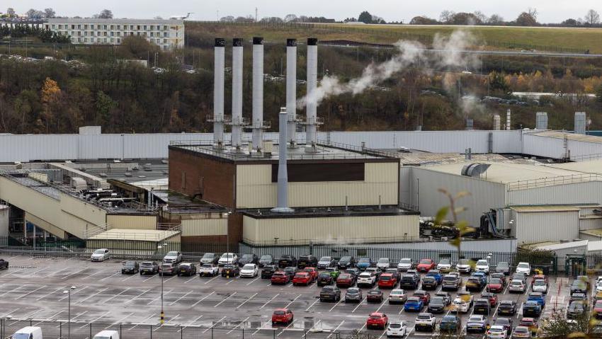 A drone shot shows the scale of the Vauxhall factory in Luton. In the centre is the plant itself with smoke coming from a large flue on the roof. There is a busy car park in the foreground and the runway of Luton Airport, with a plan on it, can be seen on the horizon.