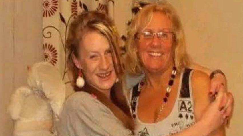 Sabrina Lyttle smiles while embracing her mother Christine Lyttle, who has shoulder length blonde hair and glasses, around the shoulders. Sabrina is wearing red and white Christmas themed earings and a grey top. 