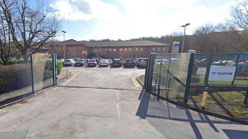 Open green gates and metal fence next to a white sign saying "Teledyne". Behind is a car park full of cars and beyond is a low brown two-story building.