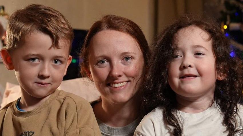 Youngsters Theo and Evie are sitting on their mother Chloe Green's lap and smiling brightly at the camera. There is a lit Christmas tree behind them.