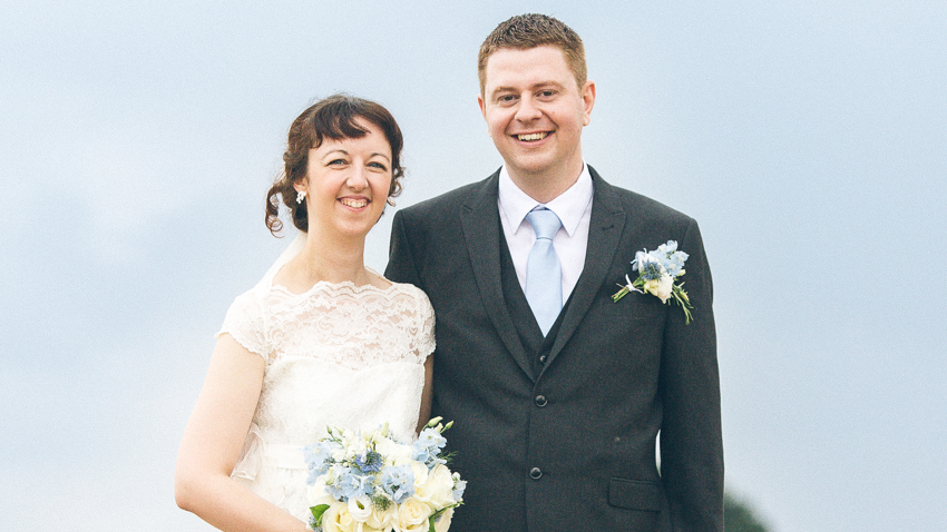 A brunette woman is wearing a wedding dress and carrying a bouquet. A sandy haired man is wearing a grey three-piece suit and pale blue tie with a flower in his lapel. Both are smiling.