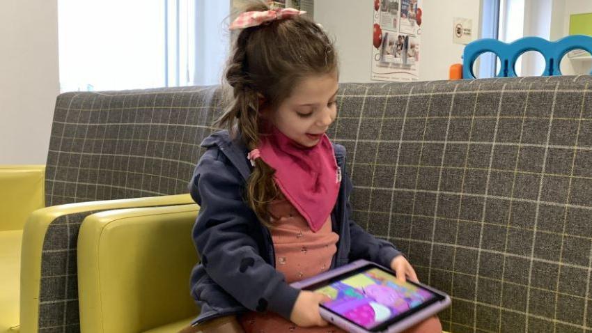 A girl kneeling on a yellow seat in a hospital ward, holding an iPad showing an episode of Peppa Pig. The girl is smiling as she looks down at the screen. She is wearing pink pyjamas, a navy hoodie and a pink bib. Her dark blonde long hair is plaited, with a pink bow at her crown and pink bobble at the end. The back of the seat is chequered white and grey and colourful stickers can be seen on white walls behind.