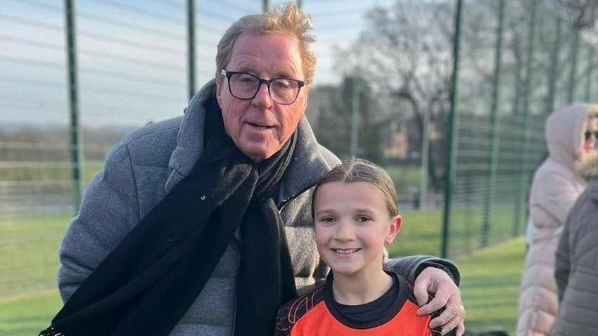 Harry Redknapp in grey jacket, black scarf and wearing square glasses. He is posing next to a blonde girl who is smiling and in football kit. 