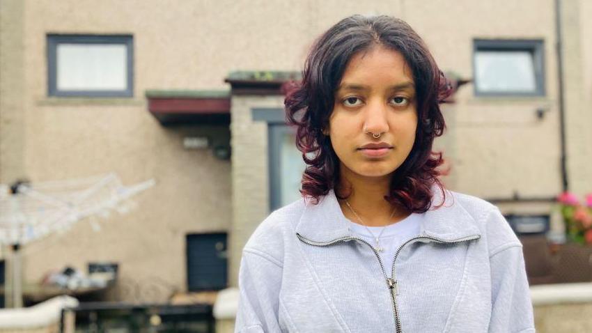 A woman, Hannah, looks forlornly into the camera. She stands in front of her home.