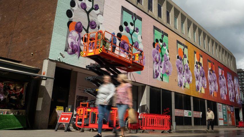 A colourful flowery mural is in the process of being painted on the side of a brick building. A stair lift is raising people up who are working on the mural. There is a street below the mural with people walking past.