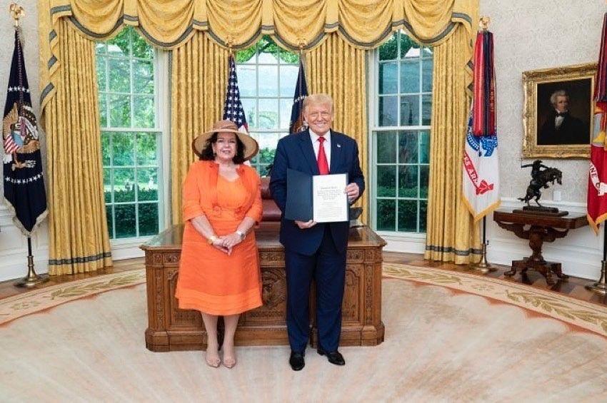 Dame Karen Pierce standing in the Oval Office with Donald Trump. She is wearing an orange outfit and a hat. Trump is wearing a suit and red tie and holding a folder towards the camera, as he smiles.