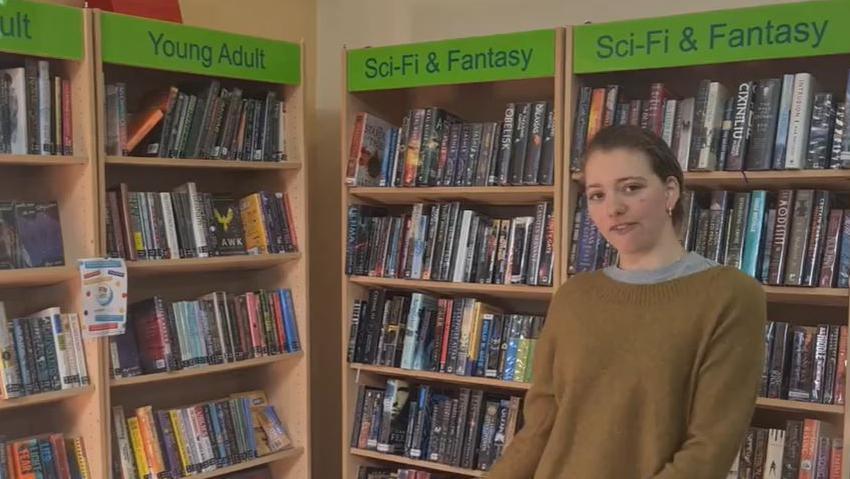 Seren wearing a light brown jumper and grey T-shirt standing in front of four bookshelves filled with books in a library