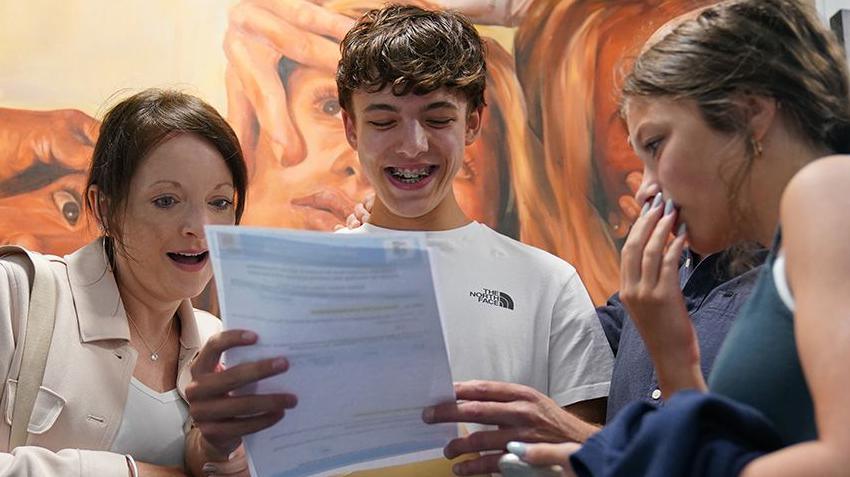 A student wearing a white North Face T-shirt smiles as he looks down at his results, printed out on a white piece of paper which he holds out in front of him. He has brown curly hair and wears braces.