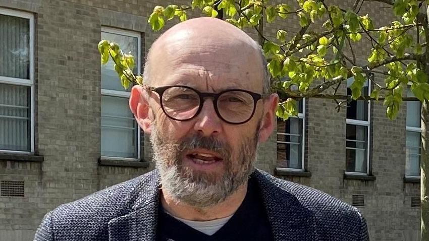 Andrew Stringer outside a block of flats. He is bald with a grey beard. He is wearing a navy jacket with a navy jumper underneath and a white T-shirt