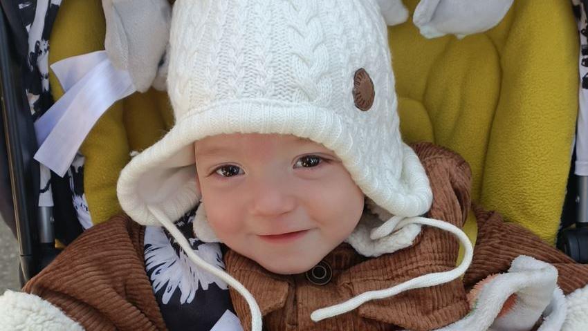 A young boy with brown eyes sits in a pushchair. He is wearing a brown jacket and white woolly hat